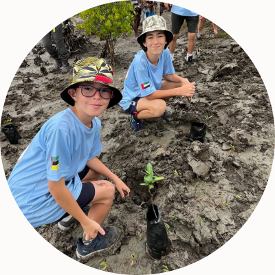 Two boys in hats and blue T-shirts kneel over a mangrove sapling they have just planted.