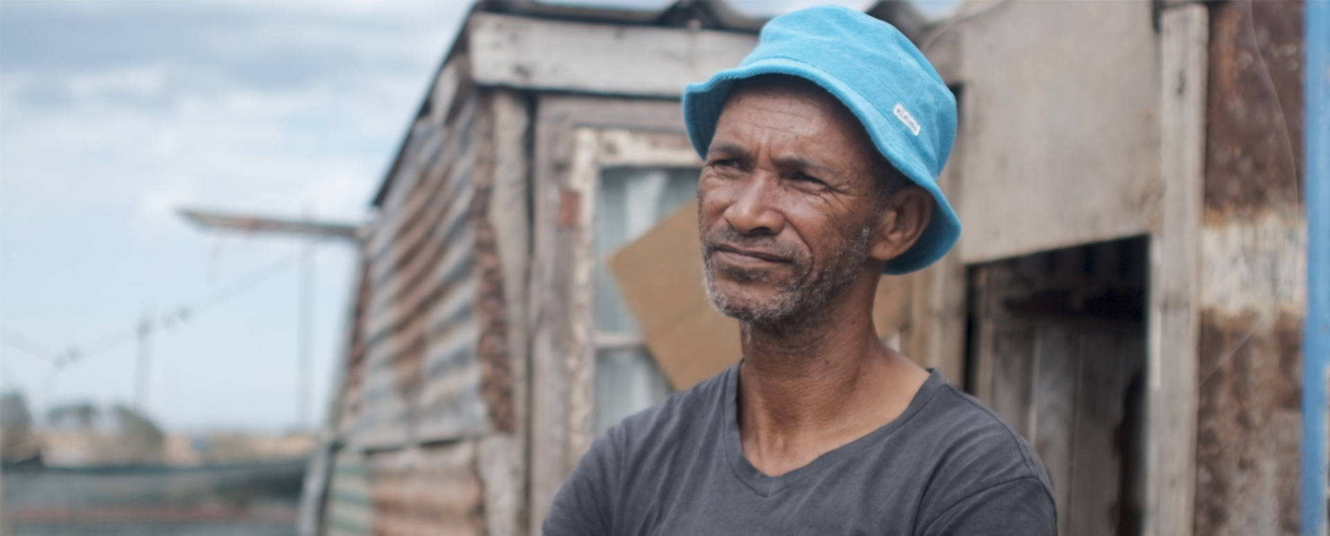 A man in a blue bucket hat and gray T-shirt stands in front of a tin-cladded building at the docks.