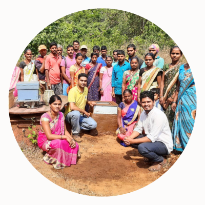 A group of people, including women in traditional Indian garb, pose around beehives.