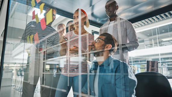 Five people gather in front of a glass wall in the office examining sticky notes in a brainstorming session. 