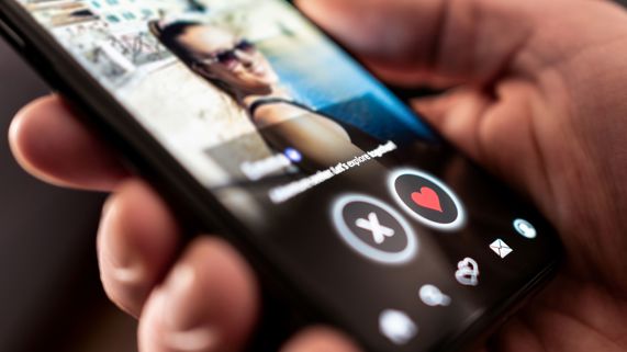 A hand holding a phone with a woman's photo on a dating app and buttons for X and a heart.