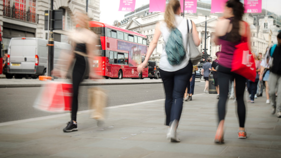 Women with shopping bags walking along a London high street with a red double-decker bus in the background. 