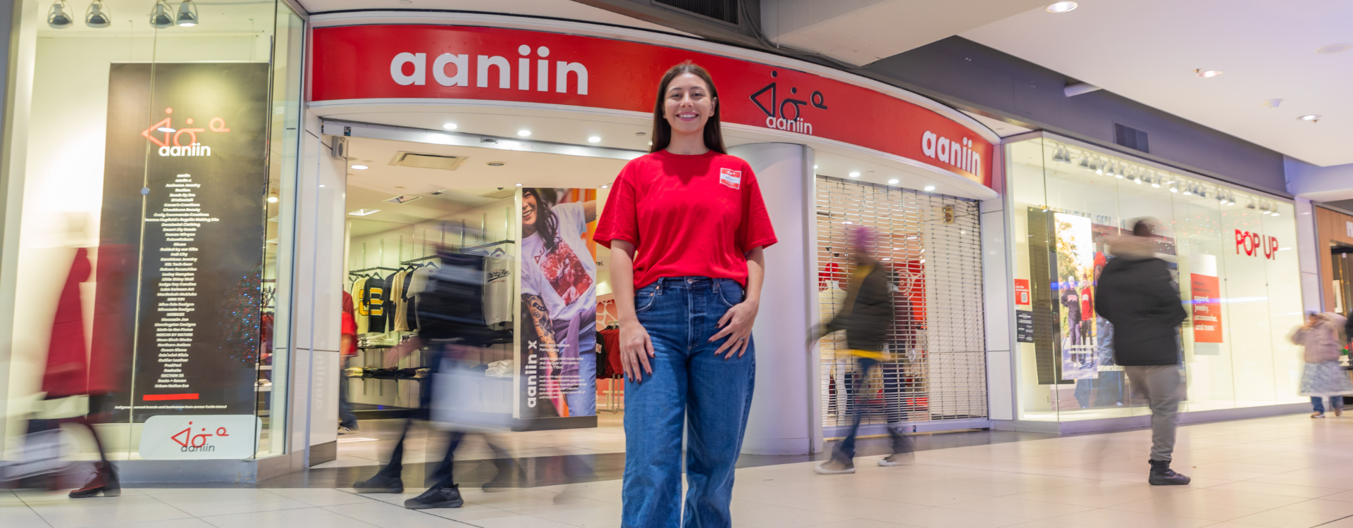 Chelsee Pettit wears a red shirt and jeans in front of her department store, Aaniin, as shoppers walk behind her. 