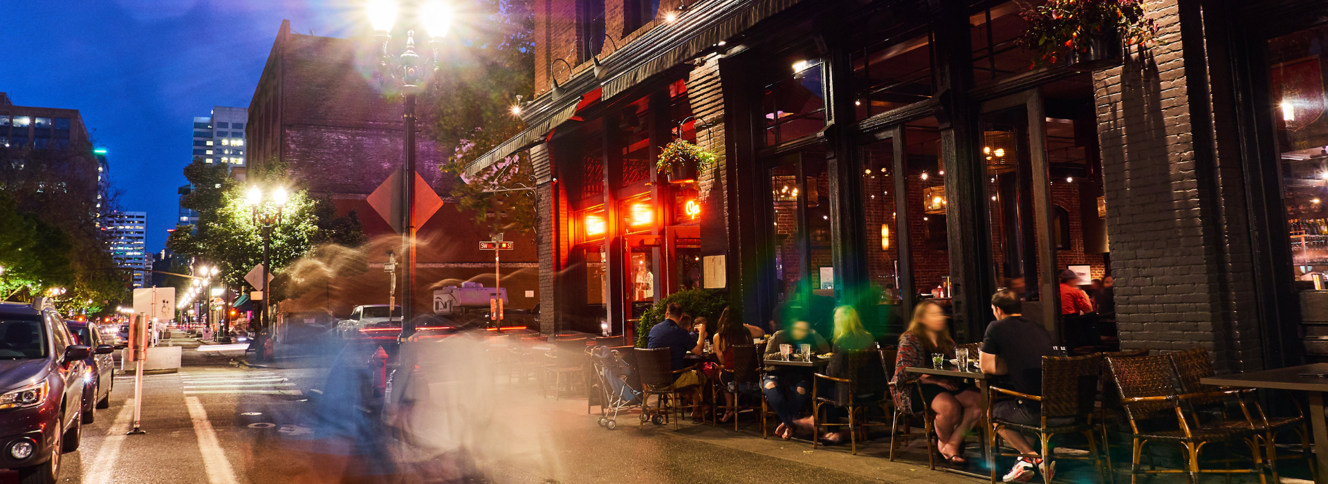 Street scene of New York City with people eating at a sidewalk restaurant at night.