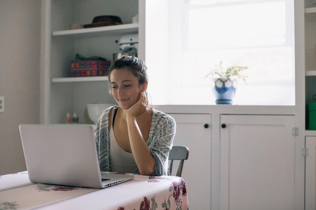 A woman sits at a kitchen table and smiles at her laptop. 