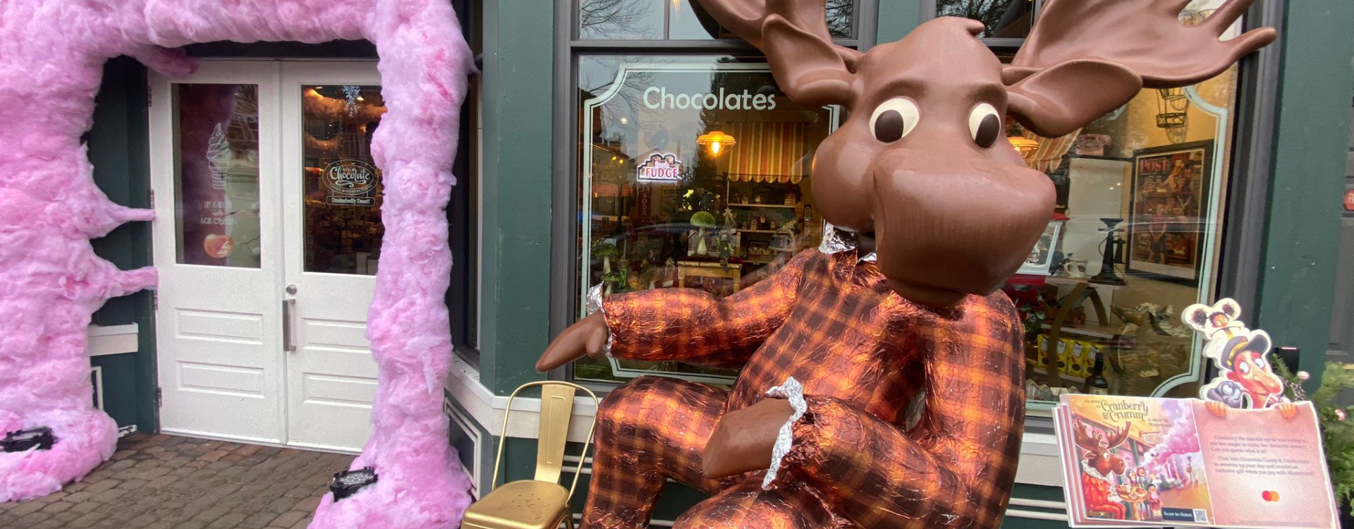 An anthropomorphic moose dressed in a suit sits in front of a chocolate shop next to Mastercard's holiday-themed storybook. 