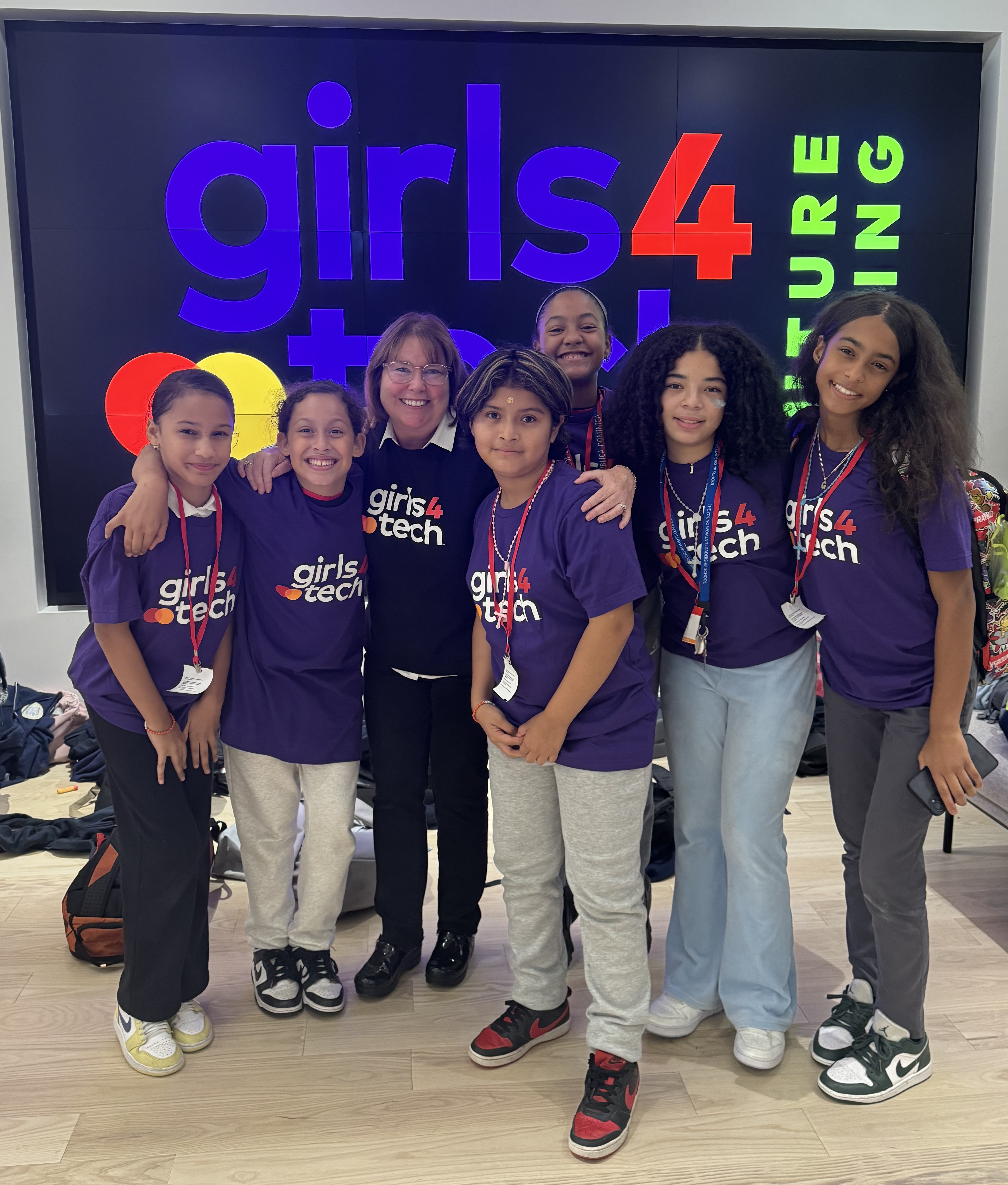 A group of Girls4Tech participants with founder Susan Warner wearing Girls4Tech T-shirts posing in front of Girls4Tech Futurecasting sign.